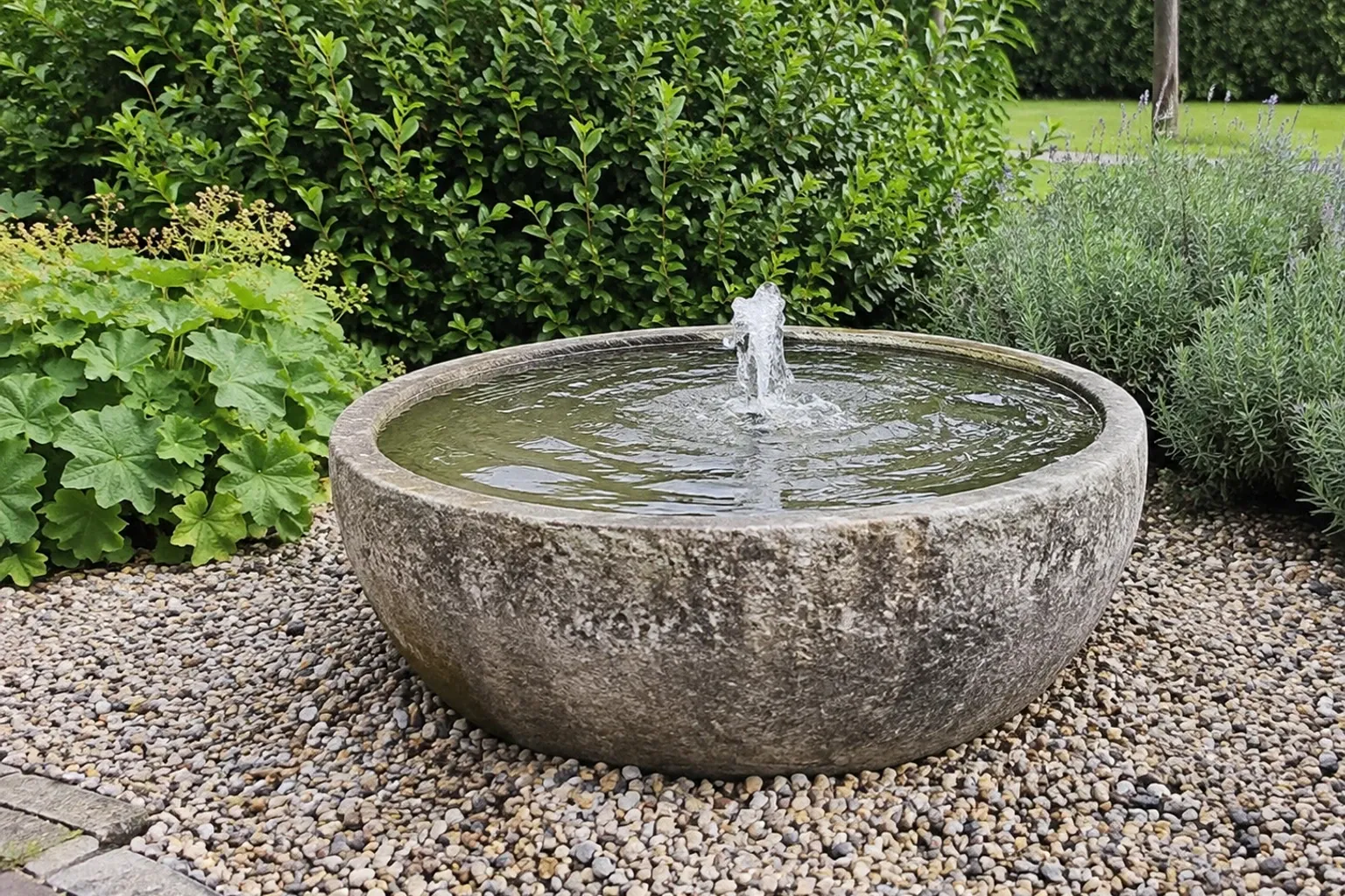 Ronde stenen tuinfontein met spuitend water, omringd door grind en groen